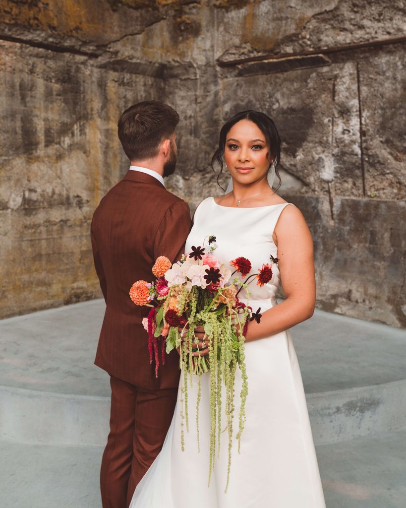 A non-traditional wedding portrait of a bride and groom. The bride is facing the camera with a more serious facial expression while the groom is liking arms with her and looking the opposite direction away from the camera. The bride is wearing a modern white wedding dress and holding a very colorful cascading bouquet of flowers. The groom is wearing a chocolate brown suit. Behind them is a rustic concrete wall at The Ruins wedding venue in Hood River.