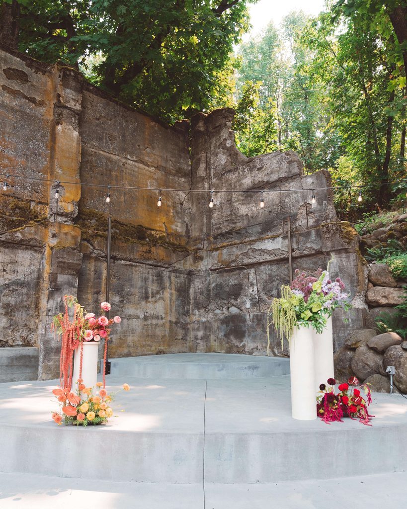 Colorful wedding ceremony flowers sitting on cream cylinder pedestals on a curved concrete stage at The Ruins, Hood River. A rustic concrete wall and large trees are in the background.