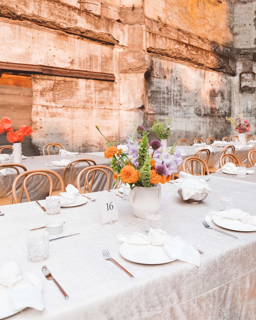 A bright, colorful floral centerpiece sitting on top of a large rectangular table with a beige velvet tablecloth. Light wooden bentwood chairs are along the table. The backdrop is a rustic concrete wall at The Ruins wedding venue in Hood River, Oregon.