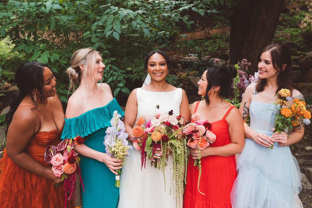 A photo of a bride and her bridesmaids. The bride is smiling at the camera and her bridesmaids are smiling at her. They are all wearing dresses of different colors (orange, teal, white, red, and light blue) and holding bouquets of flowers in bright colors.