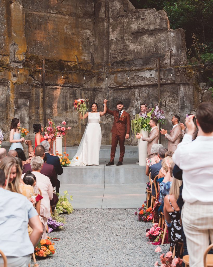 Wedding ceremony at The Ruins in Hood River, Oregon. The couple is the central focus with hands raised and cheering. The backs of their guests are shown and they are cheering and celebrating the couple. The venue is rustic with partially broken down concrete walls, which is contrasted by the brightly colored, flowers along the aisle and at the front of the ceremony space.