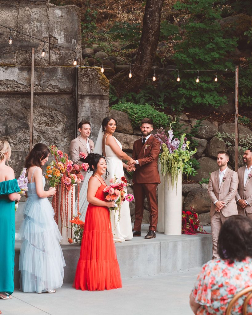 A wedding ceremony photo at The Ruins in Hood River, OR. From the perspective of the guests looking at the couple on a concrete stage with their wedding party standing at the base of the stage. The stage is decorated with brightly colored wedding flowers sitting atop cream cylinder pedestals.