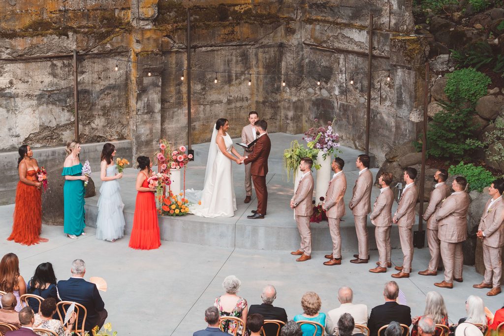 An above view of a wedding ceremony at The Ruins wedding venue in Hood River, Oregon. The couple is standing on a concrete curved stage with a rustic concrete wall behind them. On the stage, there are bright, colorful floral arrangements on cream cylinder pedestals. Just below the stage is the full wedding party. The bridesmaids are wearing brightly colored dresses with each one being unique. The groomsmen are wearing tan suits and brown shoes.