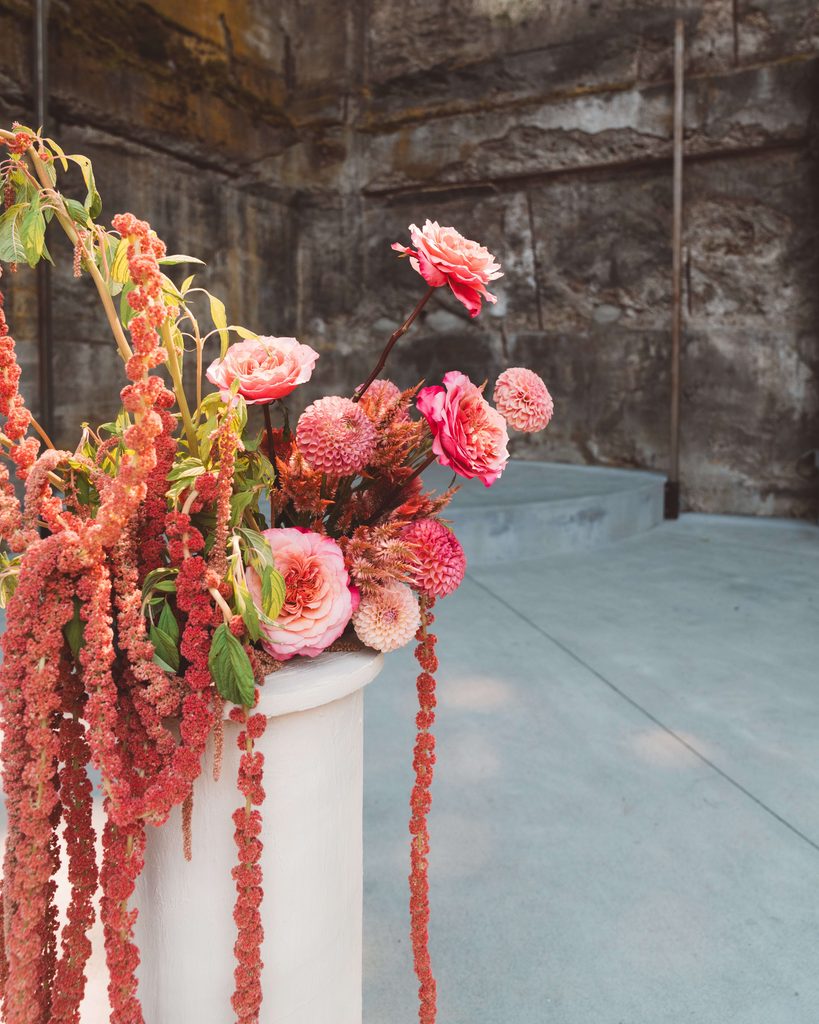 A close-up photo of a large pink and coral floral arrangement on a cream cylinder pedestal.